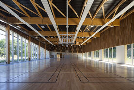 Photo de l'intérieur de la salle de sport. On voit le parquet, un panneau de basket relevé, et la vue de l'extérieur par de grandes fenêtre tout du long de la salle.