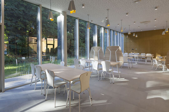 Photo de la cantine scolaire. On voit des tables rondes et des chaises avec une baie vitrée donnant sur de la verdure.