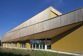 Vue de l'extérieur de la salle de sport. On voit les murs texturé en orange et le toit en bois. En premier plan de la verdure.
