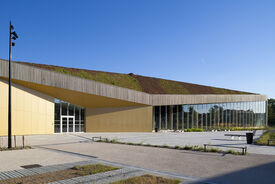 Vue de l'extérieur de la salle de sport. On voit les murs texturé en orange et le toit en bois. En premier plan le parking.