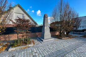 Vue en plan large du monument aux morts. Au premier plan on voit des pavés et au dernier plan une maison