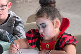 Photo où l'on voit une jeune fille déguisée et maquillée pour Halloween. Cette jeune fille est en train d'écrire, assise à une table.