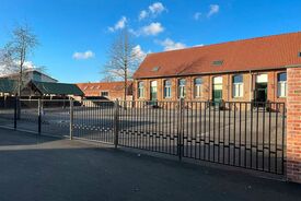 Photo de la cour de l'école primaire, vue depuis l'extérieur de l'enceinte. Au premier plan on voit les barrières séparant l'espace public et la cour de l'école.