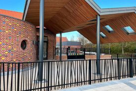 Photo de l'extérieur de l'école maternelle. Au premier plan on voit les barrières séparant le trottoir de l'école. Sur un second plan on voit un préau en bois.